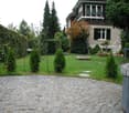 Well-maintained garden with a cobblestone pathway, trimmed hedges, a small trampoline, and a two-story house with multiple windows and shutters in the background.