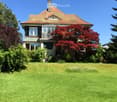 Two-story house with red-tiled roof, large windows, and a well-maintained lawn with trees and shrubs.