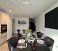 Modern dining area with a round black dining table, six dark grey chairs, a vase with flowers, and a small oven in the background.