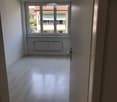 Empty room with light wooden flooring, white walls, and a large window with a view of neighboring buildings.