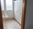 A narrow hallway with a blue carpeted floor leading to a small bathroom. The bathroom features white tiled walls, a window, two sinks, and white cabinetry.