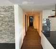 Modern hallway with a stone accent wall, wooden flooring, white cabinetry, and recessed lighting. The hallway leads to a kitchen area with stainless steel appliances and a black countertop.