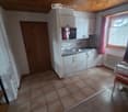 Furnished Room. The image shows a small, well-lit kitchen area with white cabinets, a sink, a stove, and a tiled floor. A wooden door is visible on the left, and a window with pink curtains is on the right. A small dining area with a chair and table is pa