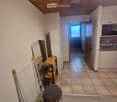 Furnished Room. A narrow hallway with tiled flooring, a small stool, a laundry rack, a small table, and a mirror. A doorway leads to another room with a visible kitchen area.