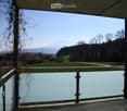 Balcony with glass railing, overlooking a green field, trees, and distant mountains.