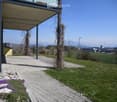 A paved pathway leads to a covered balcony area with a view of a grassy lawn, distant fields, and mountains. Small purple flowers are planted along the pathway.