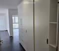 Modern hallway with sleek white cabinetry, dark wooden flooring, and recessed lighting. Features a built-in storage unit with drawers and shelves, and a door leading to a balcony area.