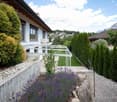 Garden area with a white house, neatly trimmed hedges, lavender plants, a paved pathway, and a view of mountains in the background.