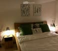 A neatly made bed with white sheets, a headboard, a nightstand with a lamp on both sides, two picture frames above the bed, a flower vase, and a small tree in a vase