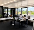 Modern office space with large windows, a desk with multiple monitors, a laptop, and a comfortable chair. The room features a sleek design with a black carpet, white walls, and a view of greenery outside.