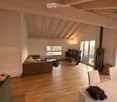Open-concept living and dining area with wooden flooring, exposed wooden beams, large windows, and a fireplace. Features a sofa, coffee table, TV, dining table with chairs, and a backpack on the table.