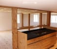 An unfurnished room with exposed wooden beams and a wooden floor, featuring a black countertop and wooden cabinetry, with large windows allowing natural light to enter.
