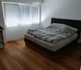 Bedroom with wooden flooring, large window with blinds, bed with dark headboard and patterned bedding, nightstand, and a shelf with folded towels.