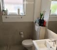 Modern bathroom with a white sink, chrome faucet, toilet, mirror, and wooden cabinet. Toiletries and a flower vase are on the cabinet. A towel rack is mounted on the wall above the toilet.