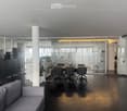 Modern office space with a grey sofa, a wooden table with chairs, a cabinet, and a potted plant. Large glass partitions and a polished black floor.
