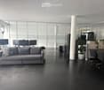 Modern, open-plan space with a grey sofa, a wooden table with chairs, multiple computer screens, a potted plant, and a fan. The room features large glass windows and a polished black floor.