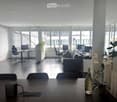 Modern office space with large windows, dark wooden floor, and white walls. Features a sofa, chairs, desks with computers, and decorative plants.