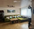 Living room with green sectional sofa, wooden coffee table, wine rack, painting, clock, and a chair near the window.