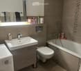 Modern bathroom with a sink, mirror, towel rack, toilet, bathtub, and showerhead. The sink has a modern faucet and a small cabinet underneath. The bathtub has a tiled border and a showerhead attached to the wall. A towel rack with towels is visible above