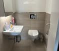 Modern bathroom with a white sink, chrome faucet, toilet, and a mirror above the sink. Beige tiles on walls and floor, with a decorative heart-shaped wall art.