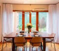 Bright dining room with large windows offering a scenic view, a wooden dining table set with plates, glasses, and a vase of flowers, surrounded by modern chairs.