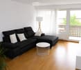 Modern living room with a black sectional sofa, white throw pillows, a white coffee table, a white TV stand, and a floor lamp. The room has wooden flooring and large windows with sheer curtains, leading to a balcony with outdoor furniture.