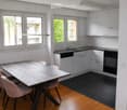Modern kitchen with white cabinets, black countertops, a wooden dining table with four pink chairs, and large windows overlooking greenery.