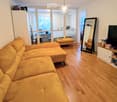Living room with a mustard-colored sectional sofa, ottoman, large mirror, TV stand, and laptop visible in the background.