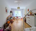 A well-lit children's bedroom with wooden flooring, a bed with a colorful blanket, a desk with a chair, a small table with a lamp, a refrigerator, and various toys scattered around, including a toy car, a toy house, and a toy stroller.