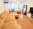 Living room with a large sectional sofa, ottoman, large mirror, television on a stand, and a small table with a chair. The room has wooden flooring and large windows with blinds.