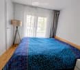 Bedroom with wooden floor, large window with sheer curtains, bed with blue patterned duvet, floor lamp, and nightstand.
