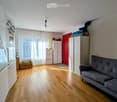 A well-lit living space with wooden flooring, a grey sofa, a refrigerator, cabinets, and a window with curtains.
