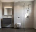 Modern bathroom with grey tiles, a dark grey vanity with a sink, a mirror above it, a toilet, and a glass-enclosed shower with a modern showerhead.