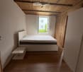 Minimalist bedroom with a white bed, nightstand, wooden floor, and exposed wooden ceiling beams. Large window with a view of greenery.
