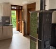 A cozy, well-lit room with a traditional wood-burning stove, light wooden cabinets, a kitchen area with a dishwasher, and a doorway leading to a green outdoor space.