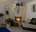 Modern living room with a white fireplace, a grey sofa with blue and green cushions, a round ottoman, potted plants, and a decorative art piece on the wall.