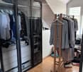 A well-organized walk-in closet with a large mirrored wardrobe, a wooden clothing rack, and a small stool. The space features wooden flooring and white walls.