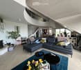 Modern living room with a large sectional sofa, coffee tables, fireplace, and large windows. The space features a mix of contemporary and cozy elements, including throw pillows, a flower vase, and a bookshelf.