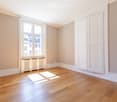 Empty room with wooden flooring, large window, radiator, and white walls.