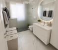 Modern bathroom with marble tiles, white sink, large round mirror, toilet, and wicker baskets.