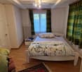 A small bedroom with a single bed, a nightstand, a chair, and a radiator. The room features wooden flooring and a window with green curtains.