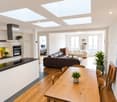 Open-plan living and dining area with modern furniture, including a dark brown sofa, a white armchair, a wooden dining table with chairs, and a sleek kitchen with white cabinets and black countertops.