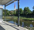 Balcony with a view of a lake, surrounded by a metal railing, featuring a covered awning and a serene landscape with trees and a church in the background.
