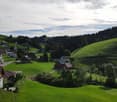 A panoramic view of a rural village nestled in green hills, with scattered houses and trees under a partly cloudy sky.