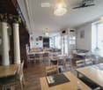 Restaurant interior with wooden tables, chairs, and place settings, featuring hanging lamps and large windows.