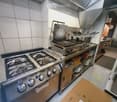 Commercial kitchen with stainless steel appliances including a gas stove, griddle, and oven. Shelves hold plates and utensils, with a cardboard box on the floor.