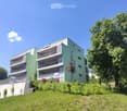 Modern multi-story apartment building with green and white exterior, multiple balconies, surrounded by trees and greenery.