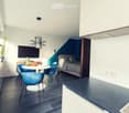 Modern, open-plan living space with a kitchen area, dining table, and a separate bedroom area. Features include a sleek black countertop, a white dining table with four blue chairs, a television, a bed with pillows and a duvet, and a stylish hanging lamp.