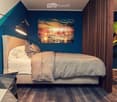 Modern bedroom with a large bed, nightstand, mirror, wall art, and a stylish lamp. The room features dark blue walls and wooden flooring.