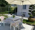 Outdoor patio area with a table, chairs, umbrella, and potted plants, surrounded by a well-maintained garden and a building in the background.